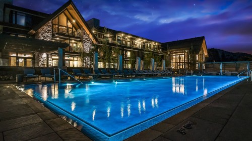 A serene outdoor pool illuminated at night, surrounded by modern buildings and lush greenery under a starry sky.