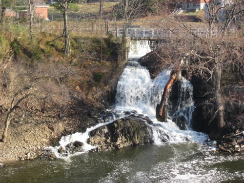 A small waterfall cascades over rocks into a calm river, surrounded by bare trees and greenery in the background.