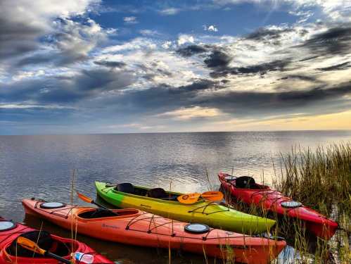 Colorful kayaks rest on the shore of a calm lake under a dramatic sky with clouds at sunset.