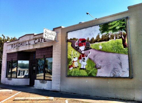 A quirky roadside cafe with a mural of cartoon animals and a truck on a grassy road. Bright blue sky above.