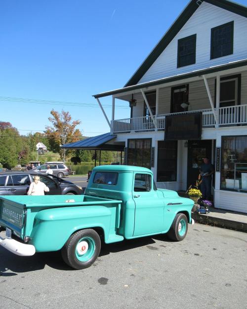 A vintage turquoise pickup truck parked in front of a white building with a porch and autumn foliage in the background.