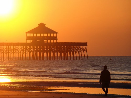 Silhouette of a person walking on the beach at sunset, with a pier in the background against a vibrant orange sky.