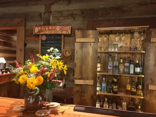A rustic bar with a wooden counter, shelves of liquor, and a vase of colorful flowers on display.