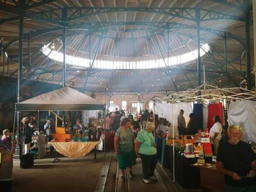 A bustling market scene indoors with vendors, shoppers, and colorful displays under a circular skylight.