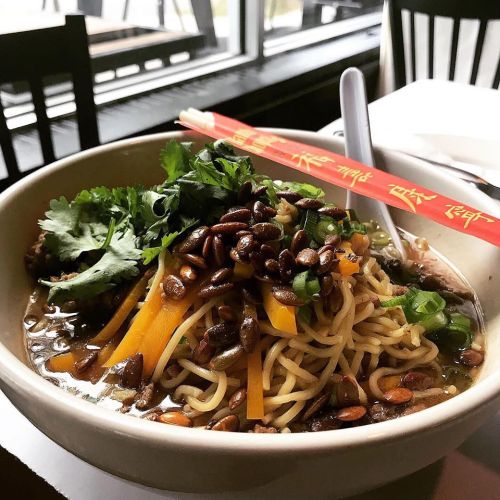 A bowl of noodles topped with cilantro, green onions, and seeds, served with chopsticks and a spoon.