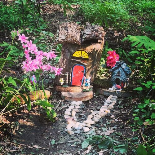 A whimsical fairy house made from a tree stump, surrounded by flowers and a stone path in a lush green setting.