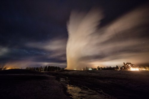 A geyser erupts against a night sky, with steam billowing and a faint glow from nearby structures.