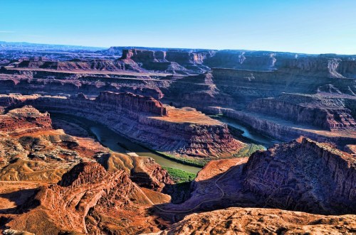 A panoramic view of a canyon with layered red rock formations and a winding river below, under a clear blue sky.