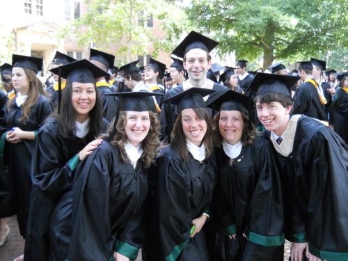 A group of six smiling graduates in caps and gowns pose together outdoors, surrounded by fellow graduates.