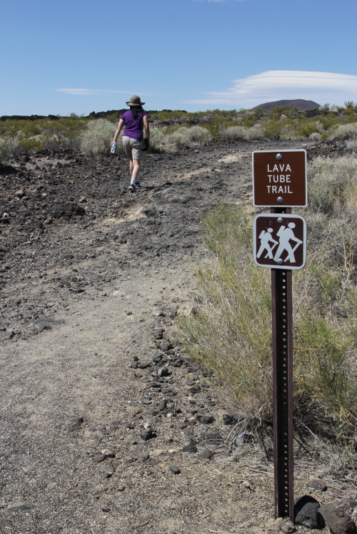 A hiker walks along a rocky path marked by a "Lava Tube Trail" sign in a desert landscape.