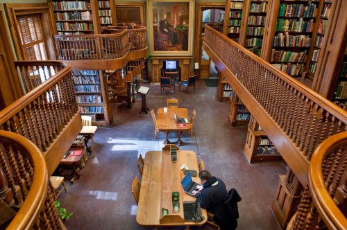 A spacious library with wooden shelves, tables, and a person working at a desk, surrounded by books.