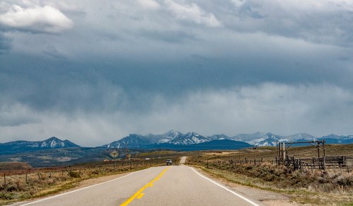 A winding road leads toward snow-capped mountains under a cloudy sky, with a fence line on the right.