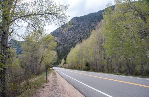 A scenic road lined with green trees, leading into a mountainous area under a cloudy sky.