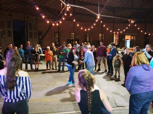 A lively gathering of people dancing and socializing under string lights in a spacious venue.