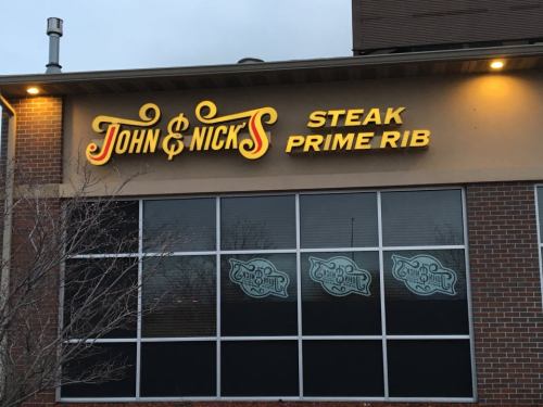 Sign for "John & Nick's Steak Prime Rib" illuminated on a brick building, with large windows displaying decorative designs.