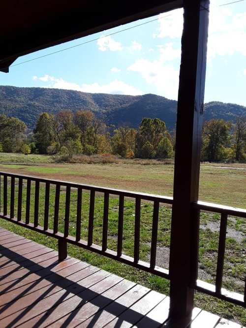 View from a porch overlooking a grassy field and mountains under a clear blue sky with scattered clouds.