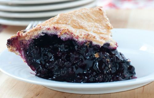 A slice of blueberry pie on a white plate, showcasing a glossy filling and a golden, flaky crust.