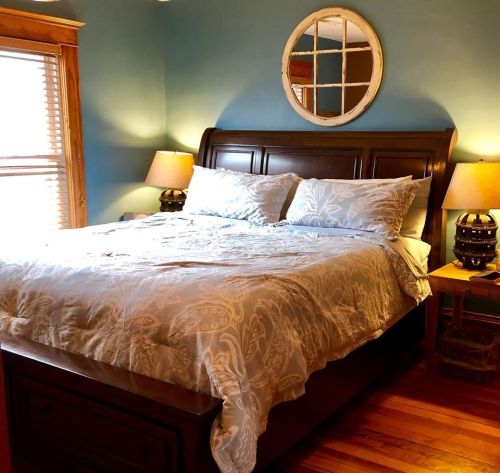 Cozy bedroom featuring a wooden bed with light blue bedding, two lamps, and a round mirror on the wall.