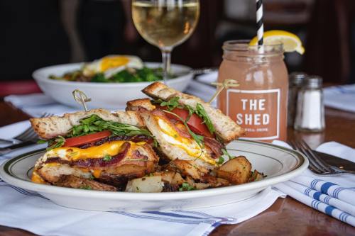 A hearty sandwich with bacon, egg, cheese, and greens, served with potatoes and a drink in a cozy dining setting.