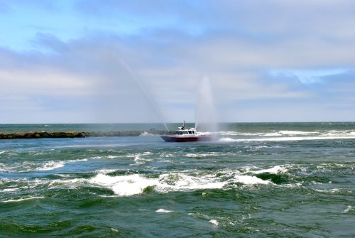 A boat sprays water as it navigates through choppy waves near a rocky shoreline under a cloudy sky.