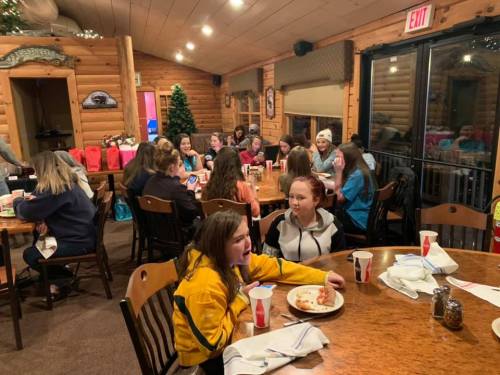 A group of young girls sitting at tables in a cozy, wooden dining area, enjoying food and chatting together.