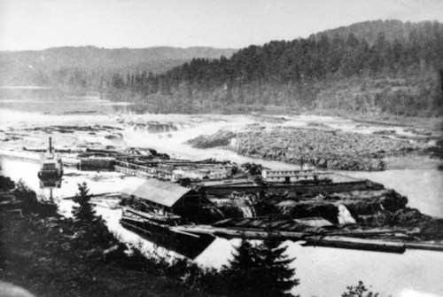 Historic black-and-white photo of a river with boats and a waterfall, surrounded by trees and mountains.