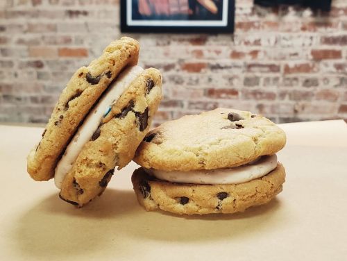 Two chocolate chip cookies sandwiching a layer of creamy filling, resting on a light brown surface.
