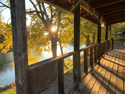A wooden porch with sunlight filtering through trees, overlooking a river surrounded by autumn foliage.