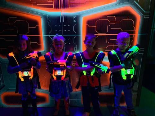 Four children in neon vests hold laser guns, standing in front of a colorful, futuristic backdrop.