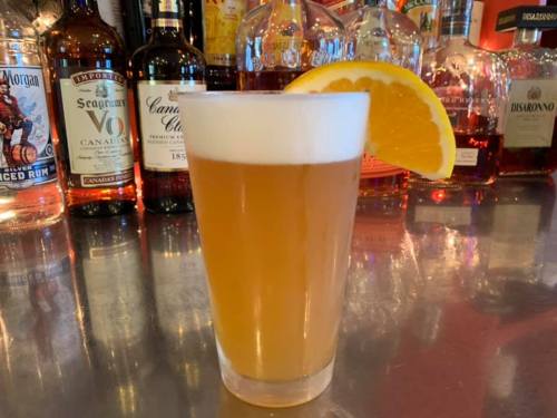 A glass of beer with a frothy top and an orange slice, set against a backdrop of various liquor bottles.