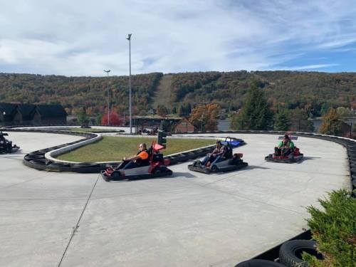 Go-karts racing on a track surrounded by trees with autumn foliage and a hill in the background.