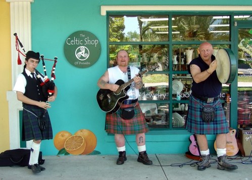 Three musicians in kilts perform outside The Celtic Shop in Dunedin, playing bagpipes, guitar, and drums.