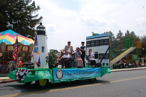 A parade float featuring Concord Point Lighthouse, with people seated and festive decorations, set against a carnival backdrop.