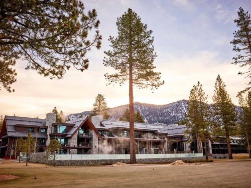 A modern lodge surrounded by tall pine trees, with mountains in the background and mist rising in the evening light.