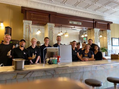 A group of smiling staff members stands behind a counter in a vintage-style diner, ready to serve customers.