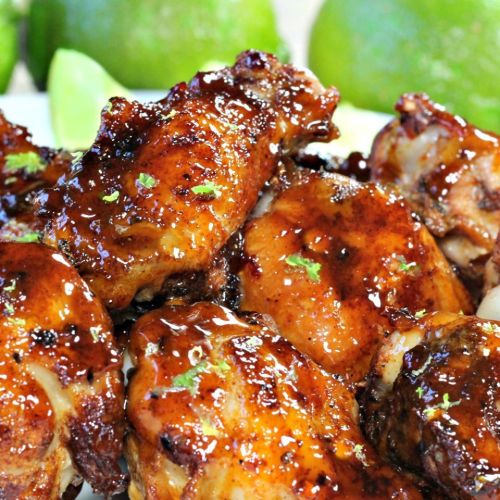 A close-up of glazed chicken wings garnished with lime zest, with fresh limes in the background.