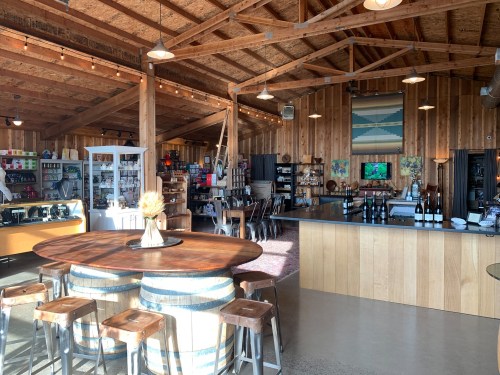 A cozy, rustic café interior with wooden beams, a large round table, and shelves filled with various products.