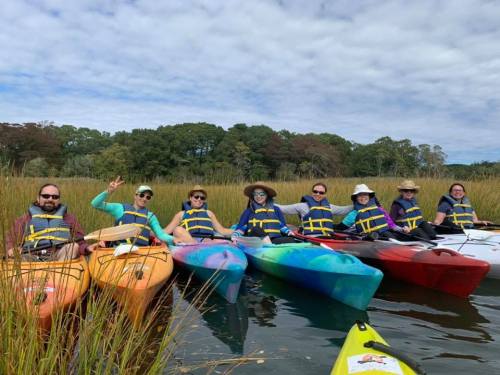 A group of nine people in colorful kayaks, wearing life jackets, enjoying a day on the water surrounded by greenery.