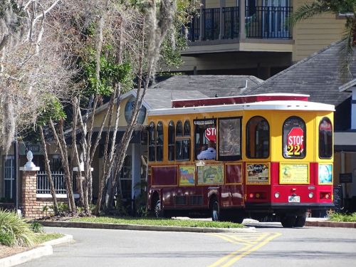 A colorful trolley bus parked near a building, surrounded by trees and a clear blue sky.
