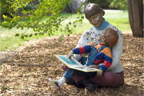A statue of a woman reading to a child, surrounded by greenery and wood chips.