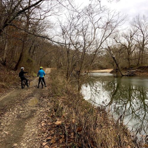 Two cyclists pause by a riverbank on a dirt path, surrounded by bare trees and autumn leaves.