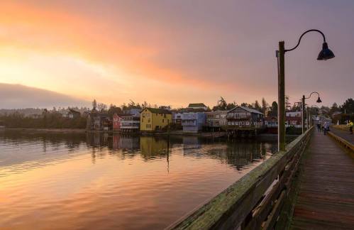 Sunset over a calm waterfront with colorful houses and a wooden pier, reflecting in the water.