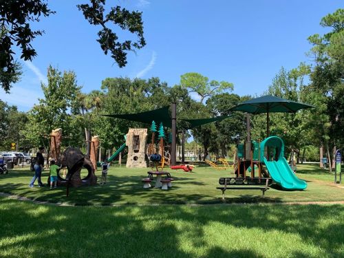 A sunny playground with slides, climbing structures, and shaded areas, surrounded by trees and children playing.