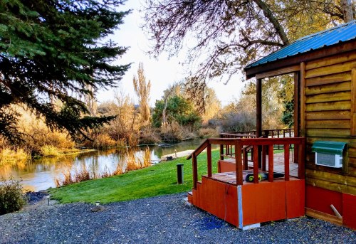 A cozy cabin by a serene river, surrounded by trees and autumn foliage, with a wooden deck and grassy area.