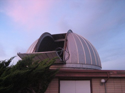 Observatory dome with an open roof against a pastel sky, revealing the telescope inside.