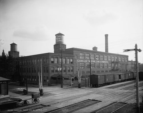 Historic Cadillac Motor Car Company building with train cars and tracks in front, set in a vintage industrial landscape.