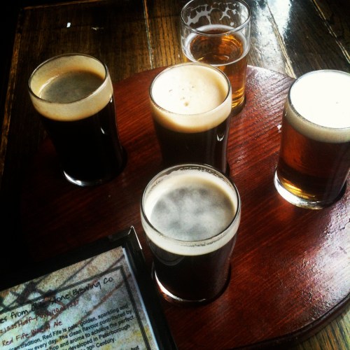 A wooden flight paddle holds five glasses of beer, with a menu partially visible on the table.