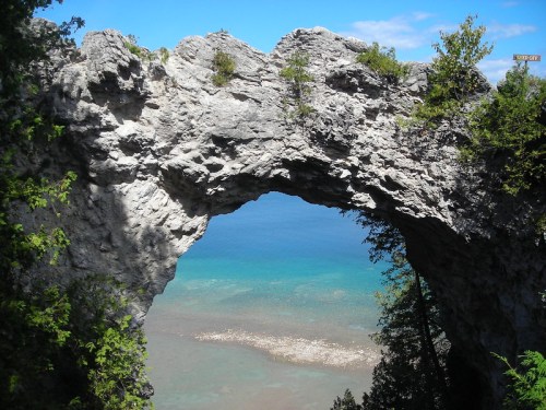 A natural stone arch frames a view of turquoise water and rocky shoreline under a clear blue sky.