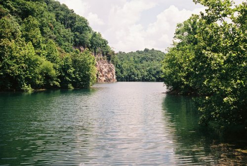 A serene lake surrounded by lush green trees and rocky cliffs under a partly cloudy sky.