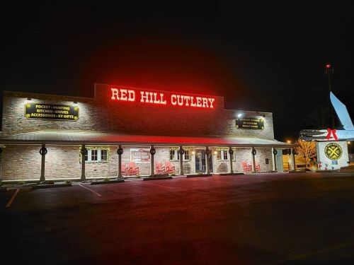 A brick building at night with a bright red sign reading "RED HILL CUTLERY" and outdoor seating.